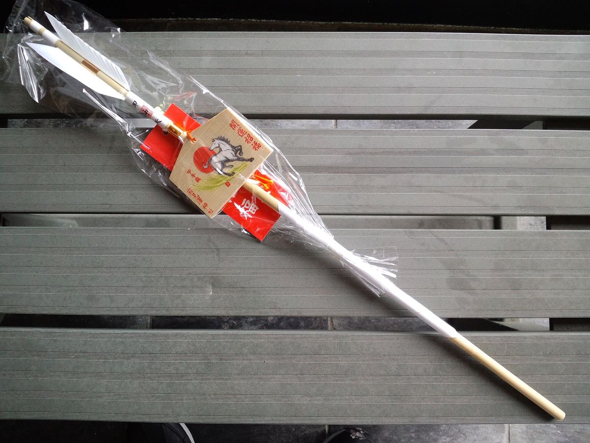 A hamaya arrow in a clear sleeve, with red paper trim and a small wooden plaque painted with the horse zodiac sign
