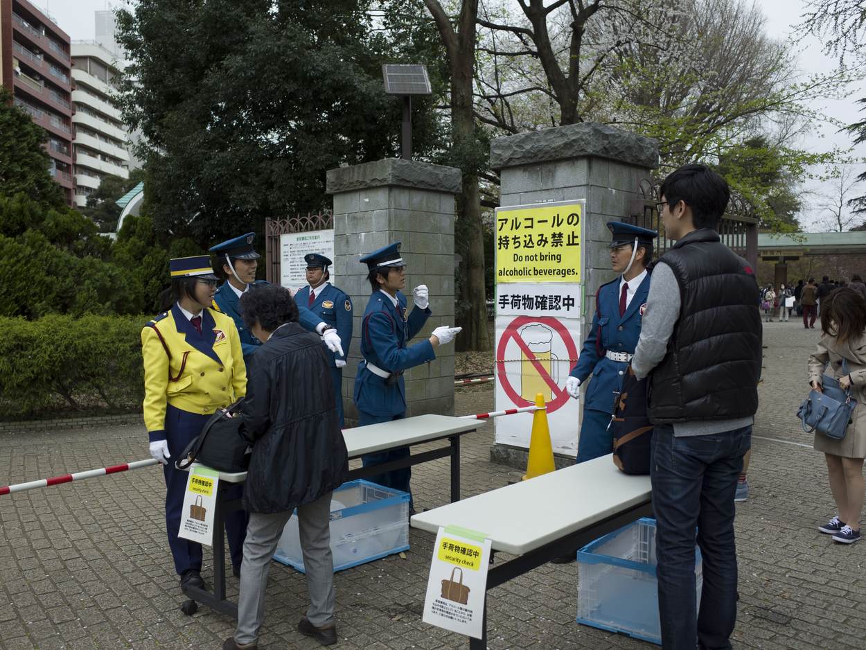 Security staff checking bags at a folding table by the entrance to Shinjuku Gyoen, next to a sign reading "Do not bring alcoholic beverages" with a crossed-out beer mug