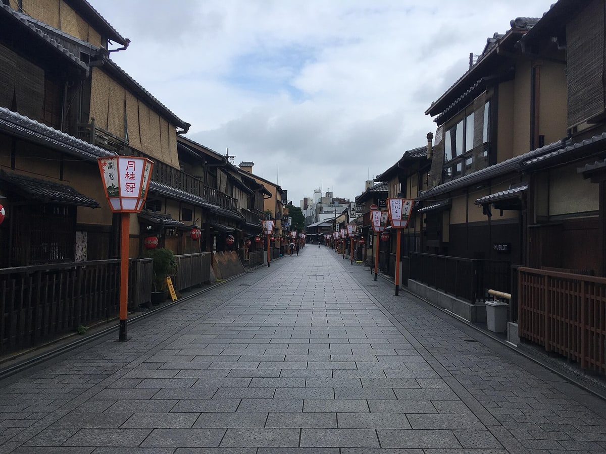 An empty Hanamikoji Street early in the morning: stone paving running the length of the frame, two-storey wooden machiya and small red lanterns on both sides, a row of orange lamp posts carrying paper shades printed with Onshukai and shop names, and only two indistinct figures far down the street.
