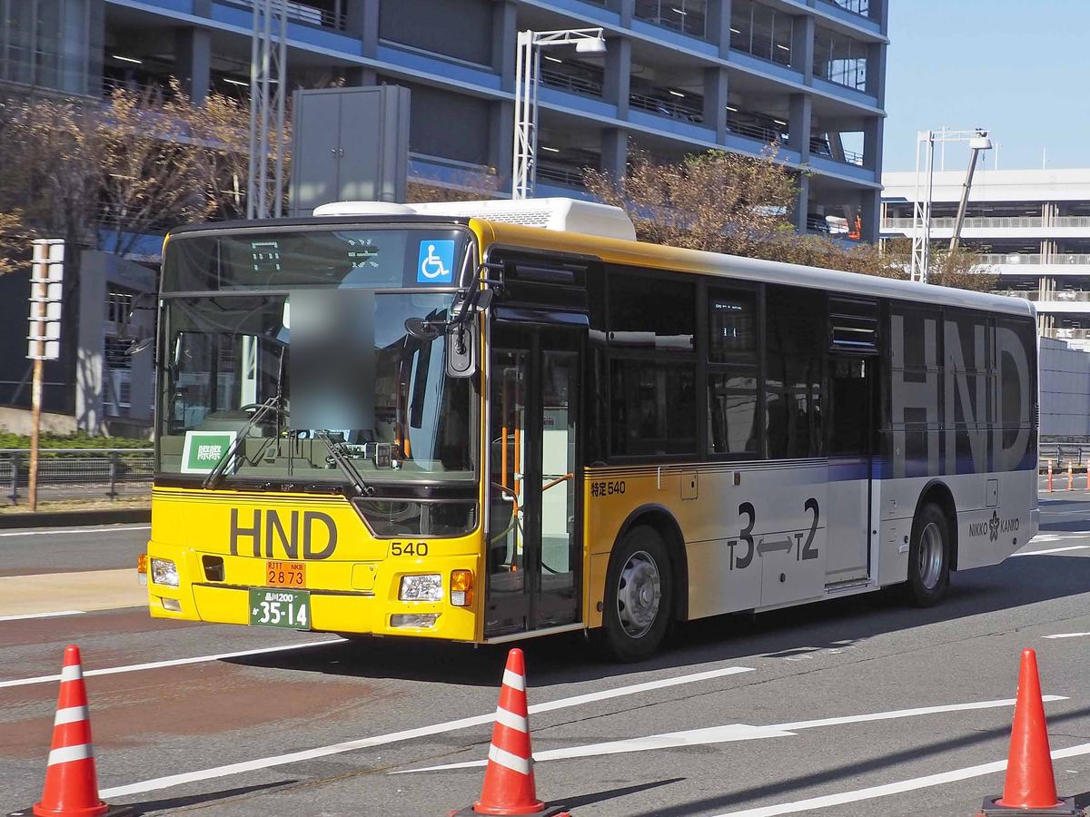 羽田機場的航廈間免費接駁巴士：黃色與銀灰色車身，車頭與車側印著大字「HND」，側面標示「T3→T2」，停在航廈前的車道上，前方擺著三角錐