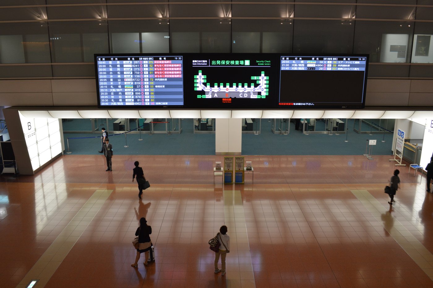 Looking down from a balcony onto the south departure floor of Haneda Airport Terminal 2. Three large screens hang side by side: blue domestic departure boards on the left and right with several lines of red text in the remarks column, and a central screen headed "Security Check" showing a gate map with a you-are-here marker and lanes A, C and D. The clock in the corner reads 19:30, and a handful of passengers stand on the wide empty floor below.