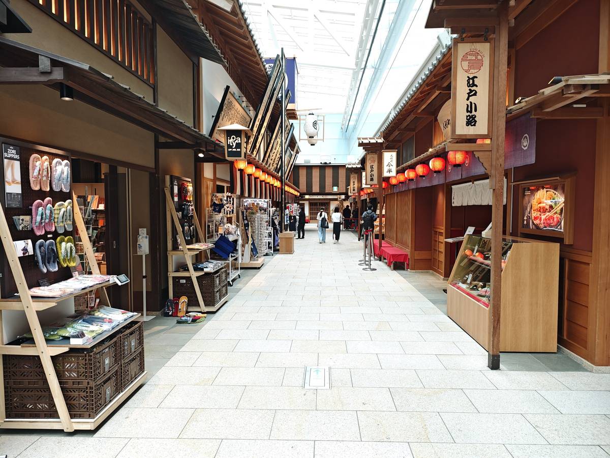 羽田第 3 航廈 4F 江戸小路:石板步道兩側是仿江戶町屋的木造店面,右邊掛著「江戸小路」直式招牌和一排紅燈籠,左邊是賣草履拖鞋的伴手禮攤,透光的天花板下只有零星幾個背影