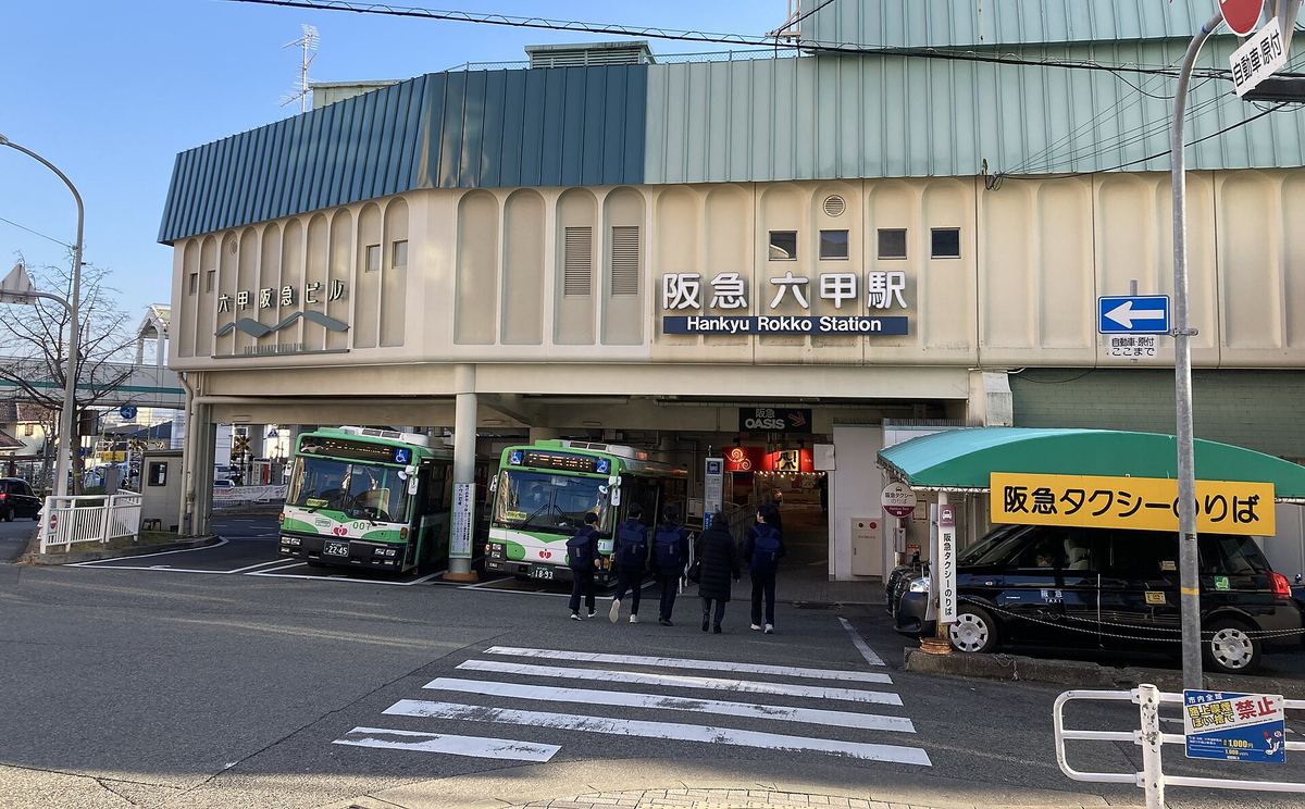 2024 年 1 月的阪急六甲站：米色站房外牆掛著「阪急 六甲駅 Hankyu Rokko Station」站名，站房樓下停著兩台綠白色的神戶市巴士，右側是掛黃色招牌的阪急計程車乘車處，幾位背著書包的學生背對鏡頭走向車站