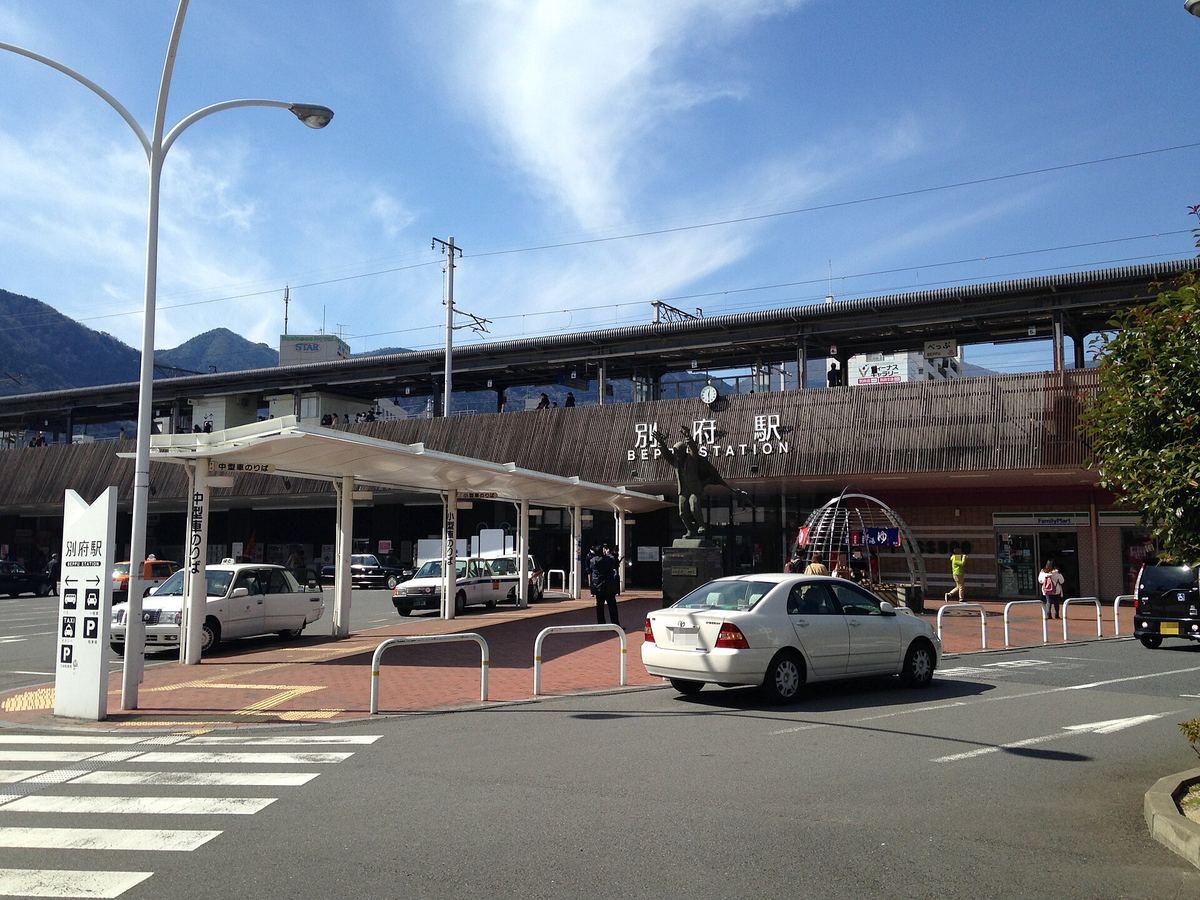 JR 別府站東口:低矮的站房外牆掛著別府駅 BEPPU STATION 的立體字,前面立著一尊高舉雙手的銅像,左邊是計程車乘車處的白色雨棚,站房上方是高架月台,背後是山