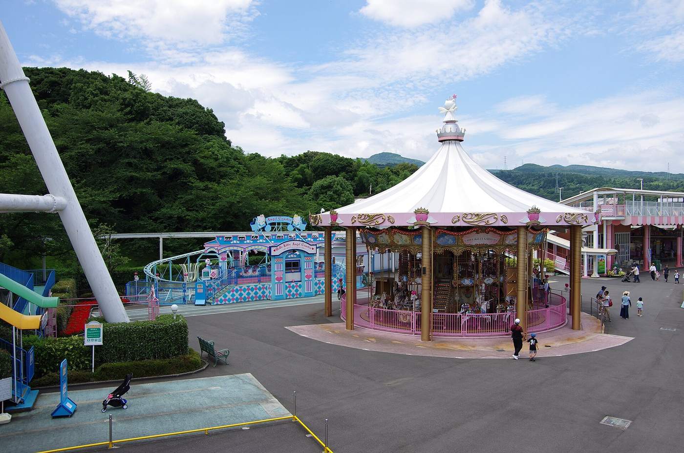 夏天陰晴天的樂園廣場:中央是白色帳篷頂、金色柱子的旋轉木馬,左邊是淺藍色的兒童設施和繞場的單軌軌道,右邊是兩層樓的粉白色建築,幾組親子在寬闊的柏油廣場上走動,背景是綠色山稜