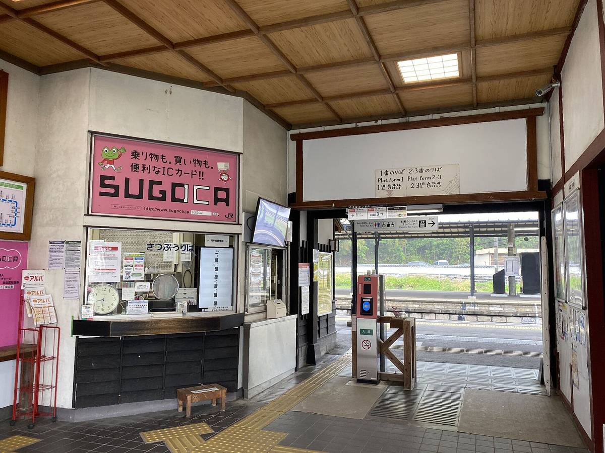 JR 杵築站的站內:木格天花板下,左邊是售票窗口和粉紅色的 SUGOCA 看板,右邊通往月台的出入口立著一根橘色的 IC 卡讀卡機,門楣上寫著「1番のりば」「2・3番のりば」,門外看得到月台與鐵軌