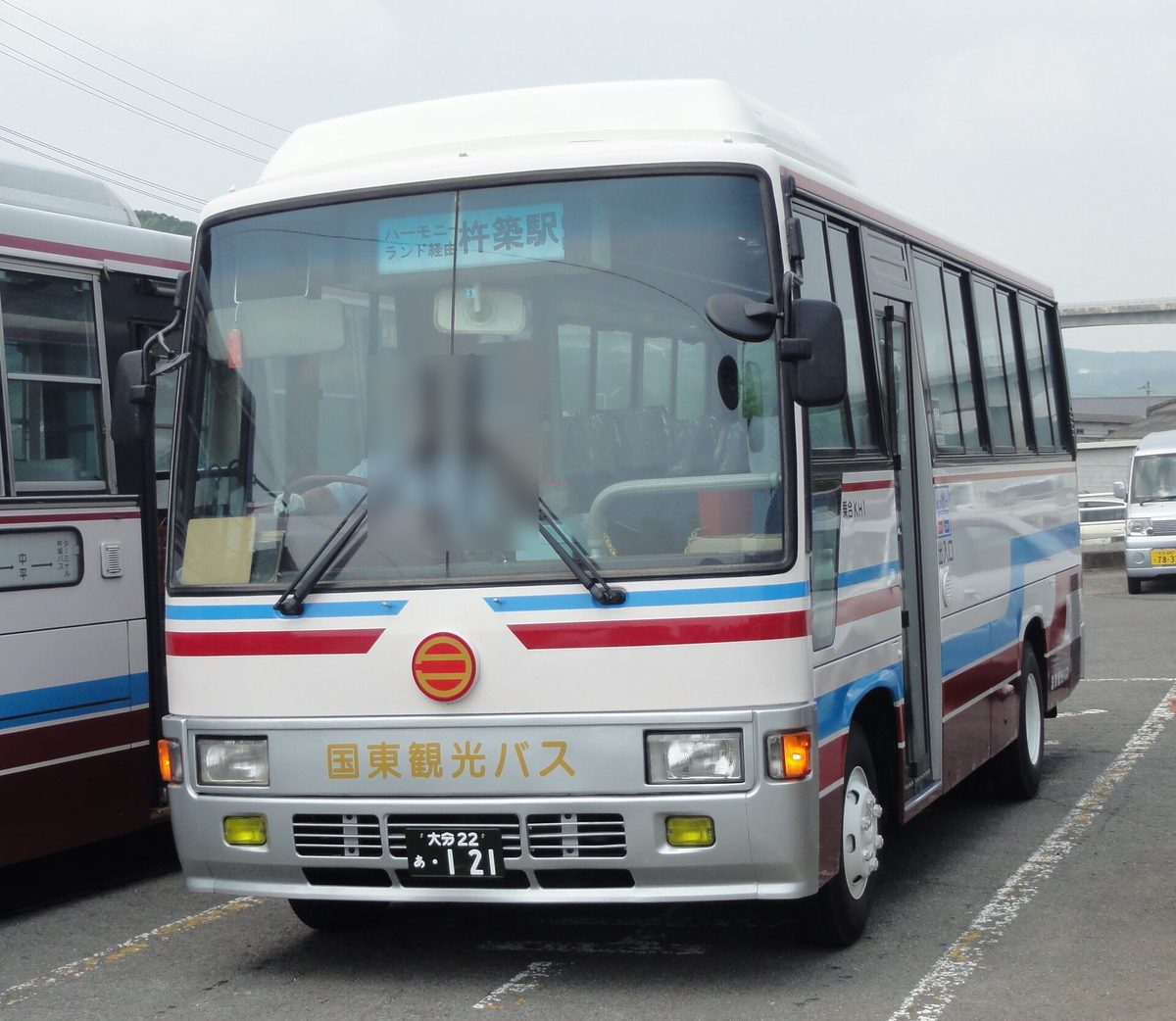 停在停車場的國東觀光巴士中型路線車,白色車身配紅藍色帶,車頭寫「国東観光バス」,擋風玻璃上方的方向牌是「ハーモニーランド経由 杵築駅」,司機座位一帶已模糊處理,左邊還停著另一台同公司的巴士