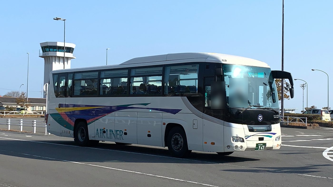 晴天的大分機場前,一台白色車身、側面有黃紫綠斜紋和「AIR LINER」字樣的大分交通遊覽車型巴士正在轉彎,左後方是白色的機場塔台,司機座位一帶已模糊處理