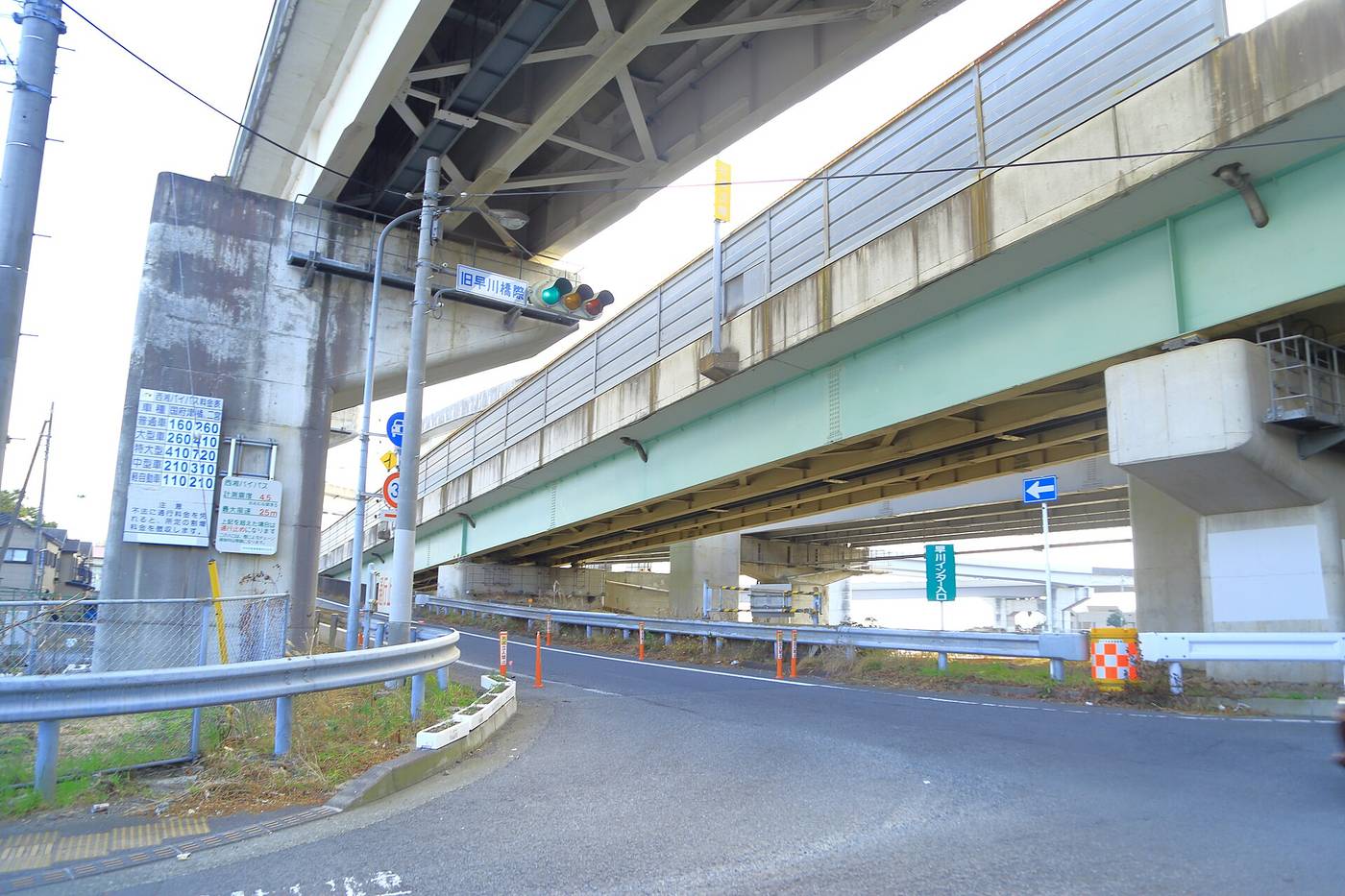 A surface road running beneath the Seisho Bypass viaduct: two levels of green steel girder deck overhead, a white toll-rate board fixed to the left pier listing five vehicle classes with their charges, a green plate reading Kyu-Hayakawa-bashi-giwa beside the traffic signal in the centre, and a green vertical sign on the right reading Hayakawa Interchange Entrance