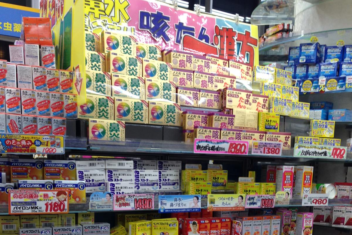 Shelves of over-the-counter medicine in a Japanese drugstore, the upper rows packed with boxes marked 鼻炎 (nasal allergy) next to cough, phlegm and cold remedies, every slot tagged with a pink or yellow yen price card.