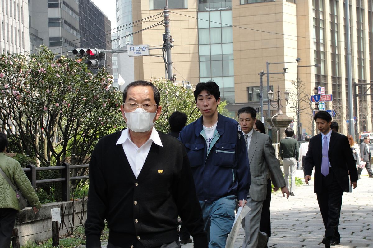 A man in glasses and a white pleated face mask walks along a sidewalk in Hibiya, central Tokyo, passing unmasked office workers, with pink blossoms on a clipped hedge to the left and a street sign reading 