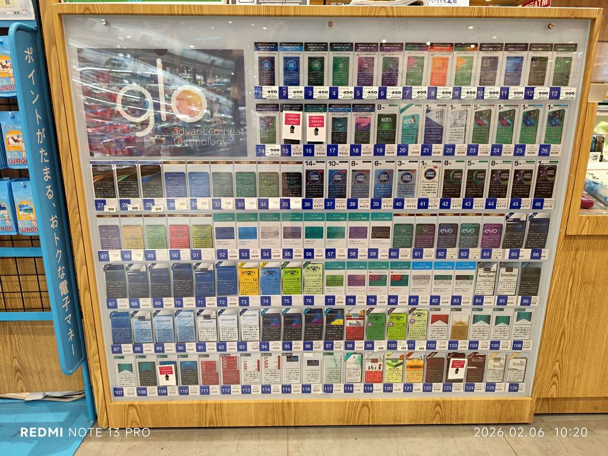 Numbered tobacco selection board behind a counter in a Japanese store, with heated-tobacco sticks along the upper rows and cigarettes below, each slot priced in yen