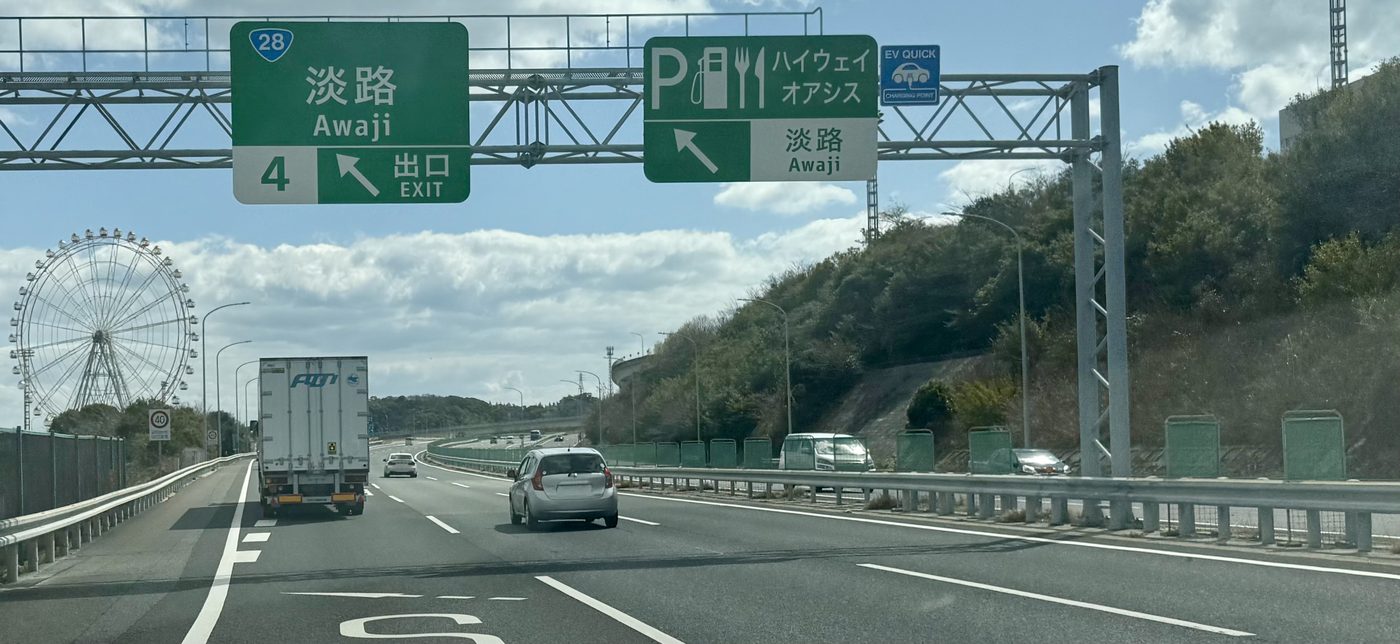 神戸淡路鳴門自動車道往德島方向的車道,上方門架掛著綠色出口標誌「4 淡路 Awaji 出口 EXIT」與「ハイウェイオアシス 淡路」,左邊遠處立著一座白色大摩天輪,右邊是長滿樹的山坡
