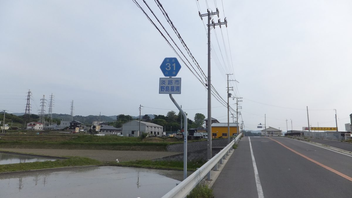 淡路島西岸的雙線道旁立著藍色路標,上面是六角形的兵庫縣道 31 號標誌,下面的地名牌寫著「淡路市 野島蟇浦」,左邊是水田和幾座電塔