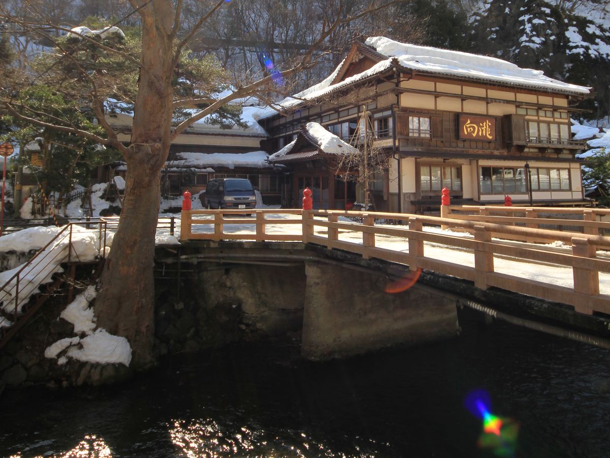 冬天白天的東山溫泉：溪流上一座木欄杆的橋，橋後是屋頂積雪的兩層木造旅館，招牌寫「向瀧」，背後山坡也有積雪