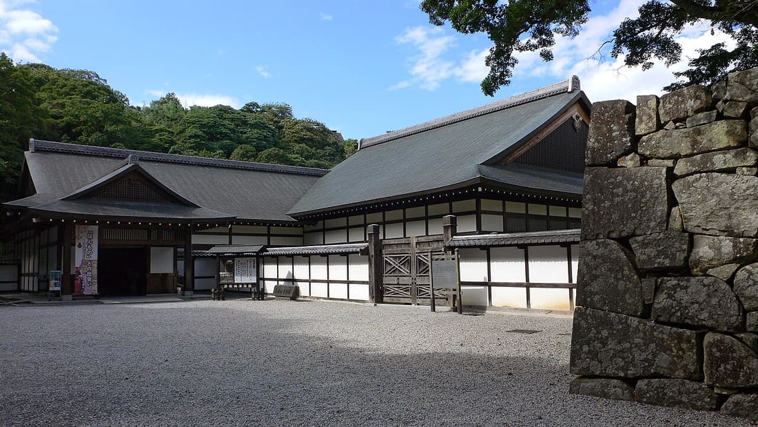 Exterior of the Hikone Castle Museum: two hipped-and-gabled timber buildings around a raked gravel forecourt, an entrance porch on the left hung with a vertical cloth banner, a long white-plaster wing with wooden lattice and a cross-braced plank gate at the centre, and a rough stone rampart in the right foreground