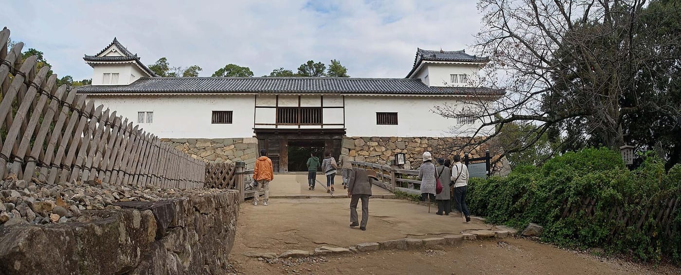 The Tenbin Yagura at Hikone Castle seen head-on: a long white-walled building on a stone base with a small black-tiled corner turret at each end, an open timber gateway beneath its centre, a wooden bridge spanning the gap in front of it, and a dozen visitors walking up the earth slope onto the bridge, with a slanted wooden palisade to the left