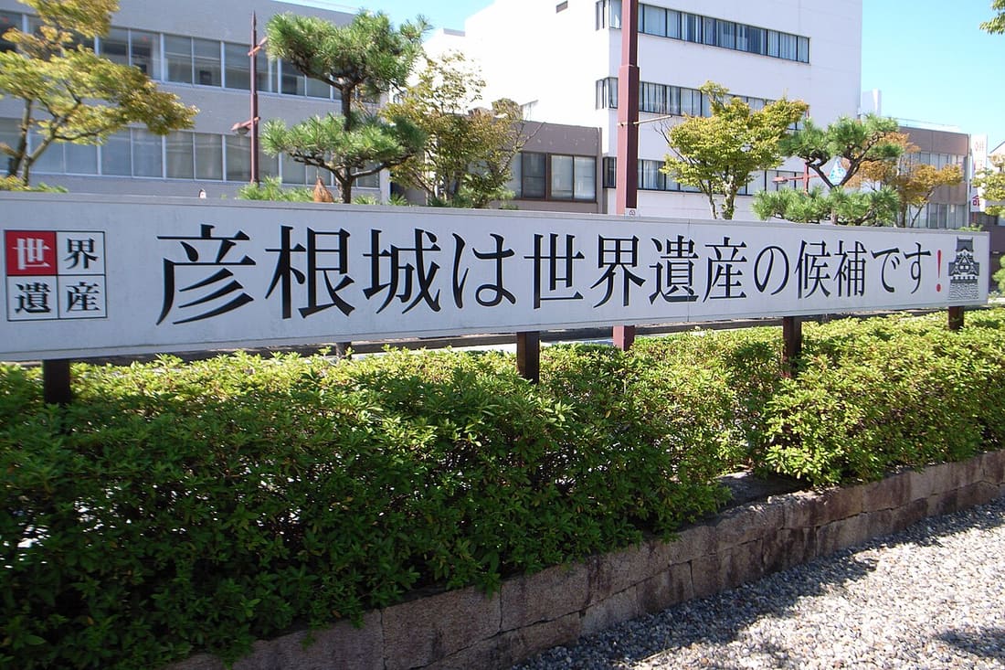 A long white roadside sign in Hikone reading, in large black Japanese characters, that Hikone Castle is a candidate for World Heritage — a boxed 'World Heritage' label at the left end, a small castle-keep pictogram at the right, standing behind a clipped hedge