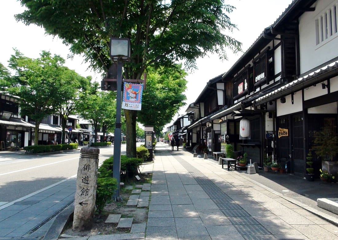 Yume-Kyobashi Castle Road in Hikone: a row of white-walled, dark-timbered machiya shophouses with lanterns and cloth signs under the eaves, a paved stone footpath, street trees and a two-lane asphalt road on the left, and a vertical banner hanging from a streetlamp