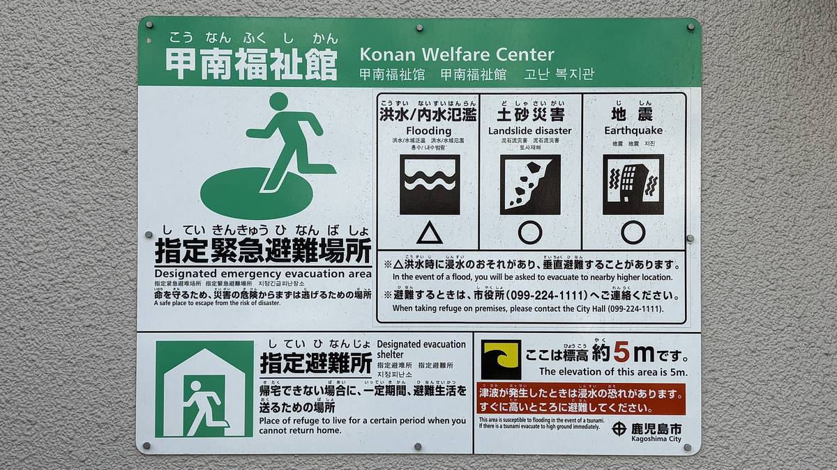 A Japanese designated emergency evacuation site signboard, green with a white running figure, with a row of hazard-type icons below
