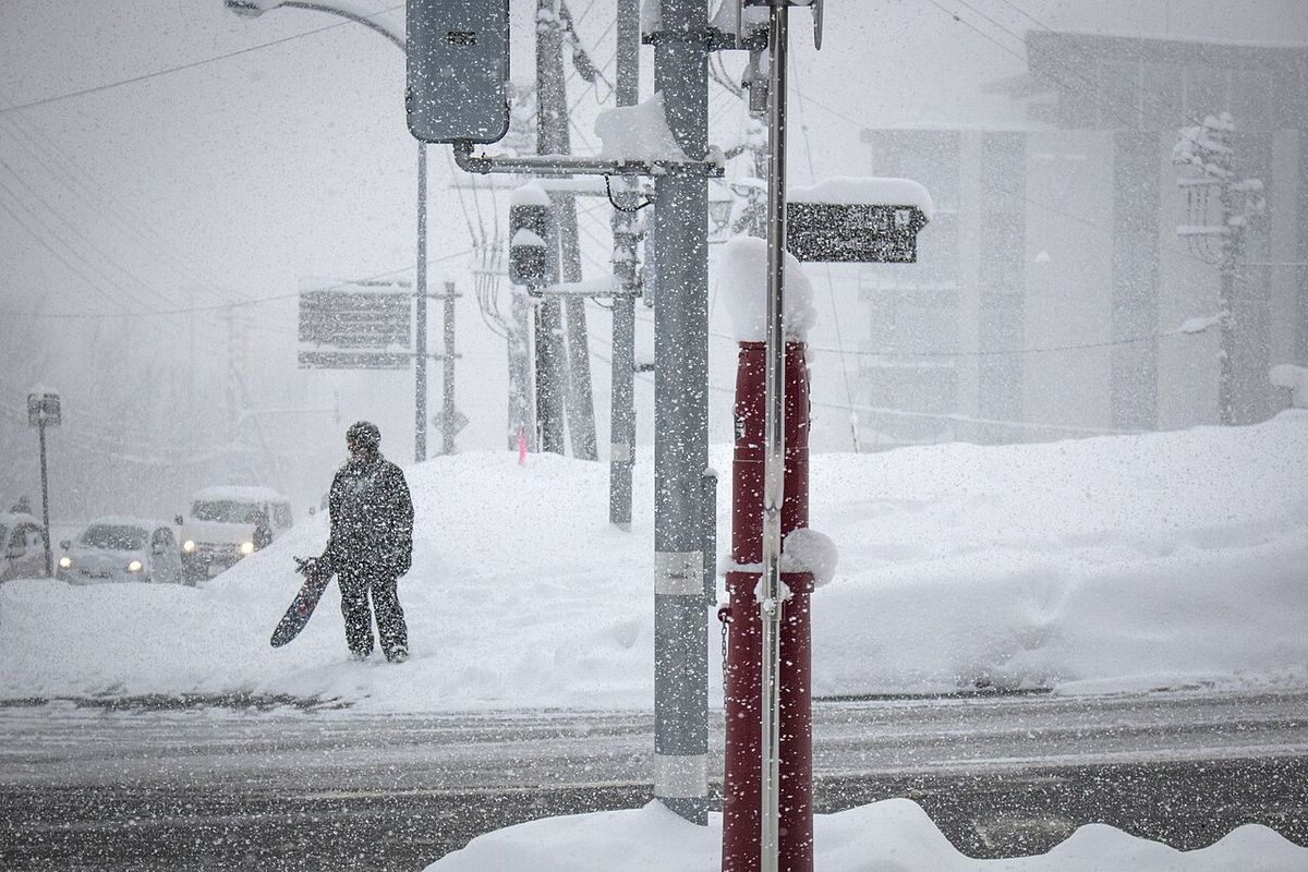 A road junction in Hirafu during heavy snowfall: traffic signal poles and red-and-white snow marker poles capped with thick snow, snowbanks taller than a person along the roadside, a snowboarder in snow gear carrying a board in the distance and cars with headlights on beyond, visibility very low