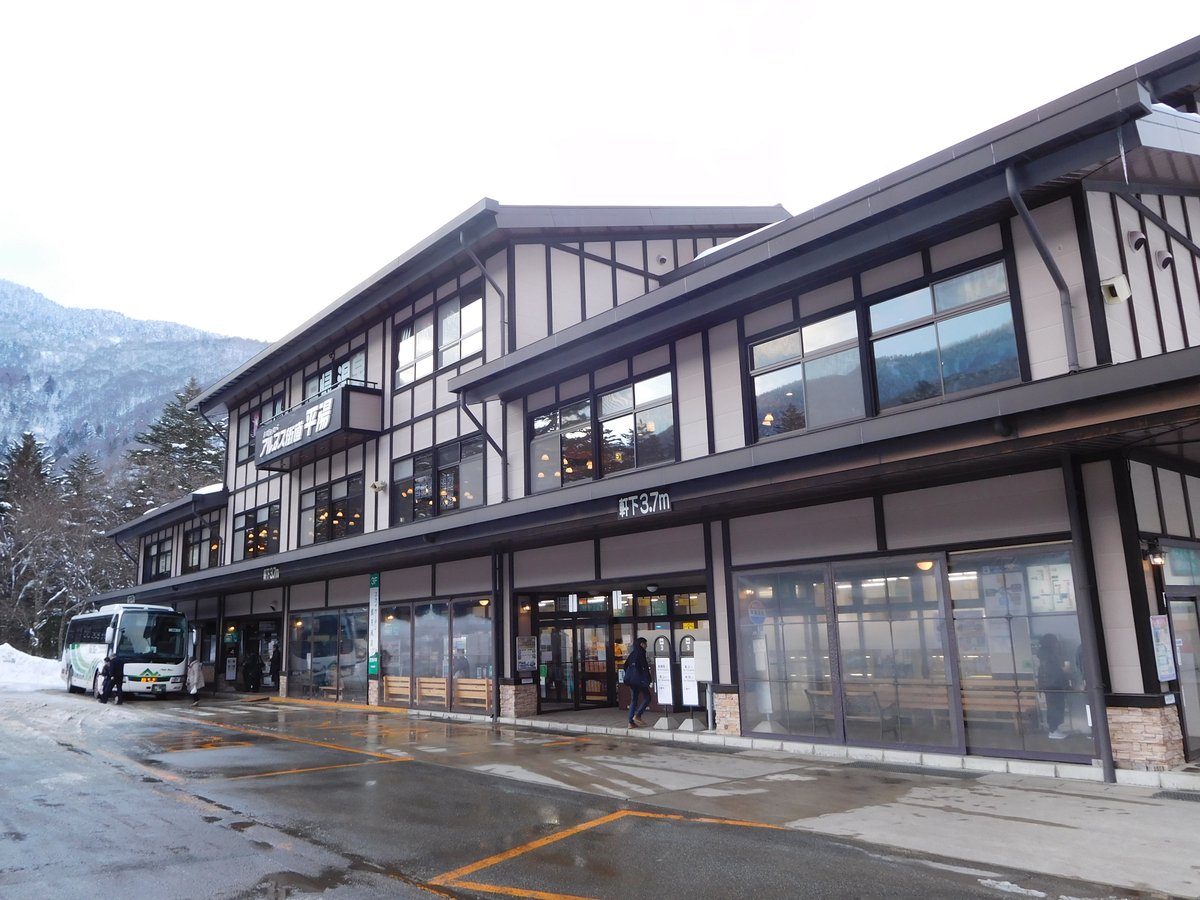 冬天的平湯巴士總站：兩層樓的木造風格站房，招牌寫「アルプス街道平湯」，一樓玻璃窗內有候車長椅，左邊停著一台白綠色的濃飛巴士，地面是融雪後的濕柏油，背後山坡積雪