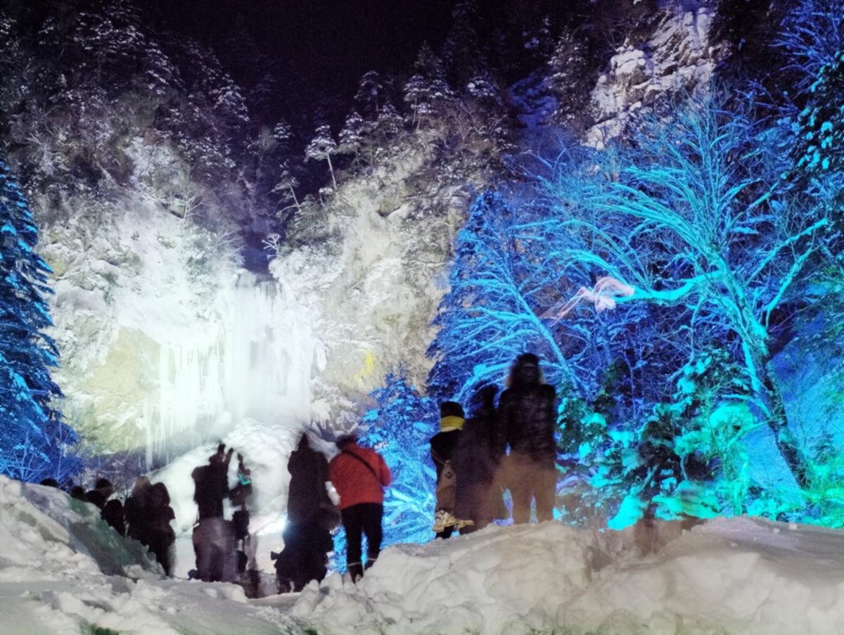 結冰祭點燈的夜晚：左邊結冰的平湯大瀑布被白光照亮，右邊雪中的樹林打上藍綠色的燈，前景十幾個穿厚外套的遊客背對鏡頭站在踩實的雪堆上拍照