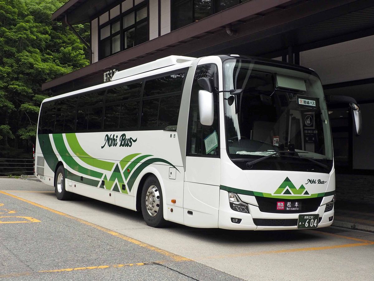 停在平湯巴士總站屋簷下的濃飛巴士高山・松本線車輛：白色車身配綠色條紋，車頭顯示「特急 松本」，背景是夏天的綠樹