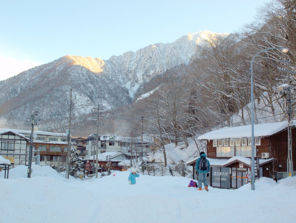 2 月的平湯溫泉：整片空地積著厚雪，右邊一間小木屋掛著「宿泊案内所」與「平湯温泉旅館組合」的字樣，一個背著背包的人背對鏡頭往那裡走，後方是幾棟旅館和山頂照到陽光的雪山
