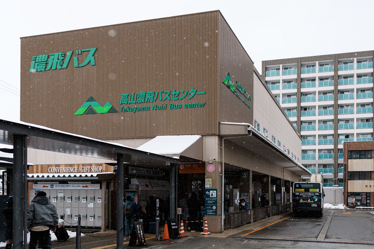 下著小雪的高山濃飛巴士中心：咖啡色外牆上寫著綠色的「濃飛バス」與「高山濃飛バスセンター」，一樓左邊是投幣式置物櫃和便利商店入口，右邊月台停著一台巴士