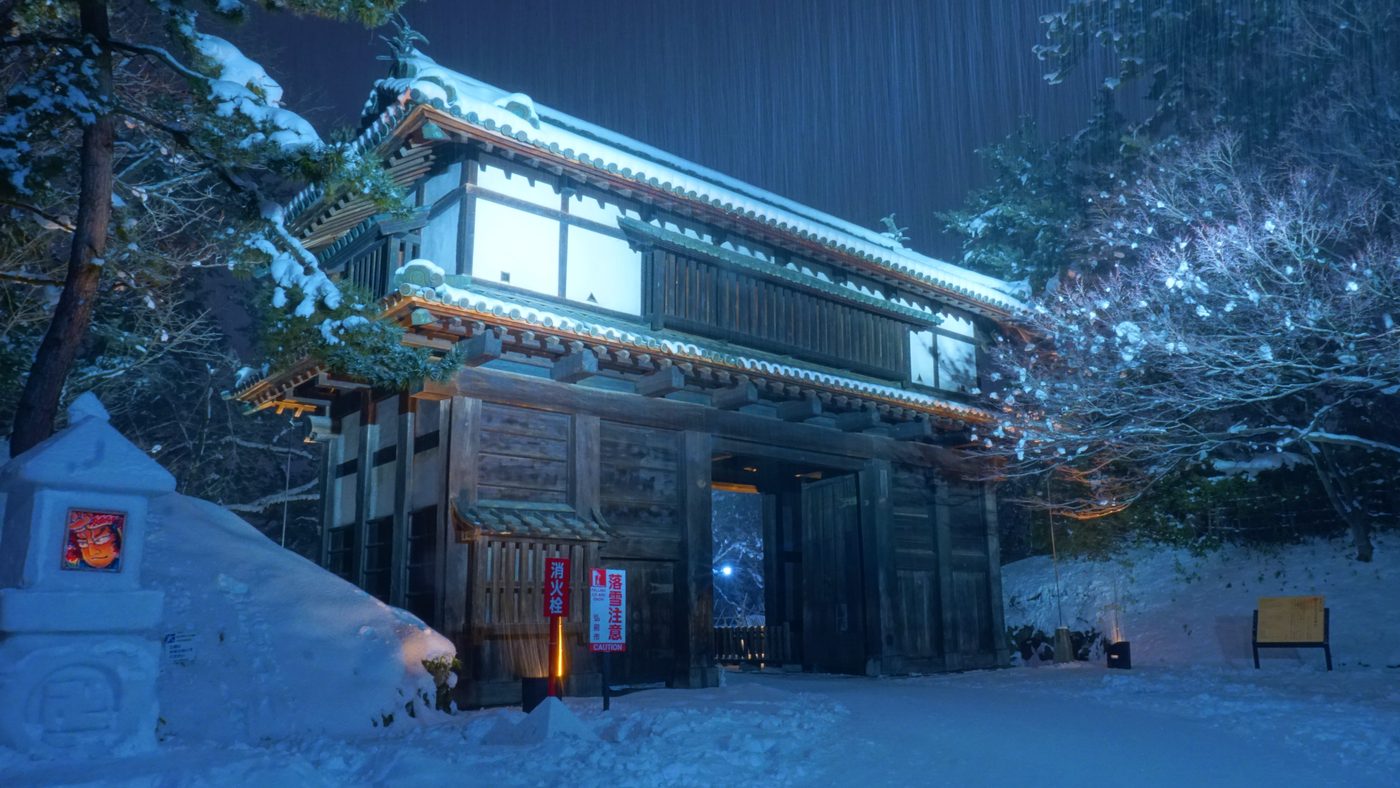 下大雪的夜裡,弘前城南內門屋頂與地面全是積雪,門前立著紅色「消火栓」與「落雪注意」的牌子,左邊一座雪燈籠的窗口嵌著一張ねぷた風格的紅色人臉畫