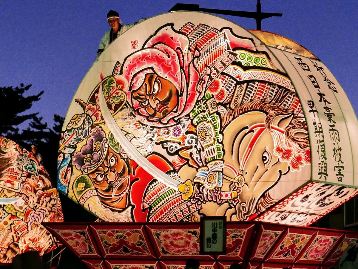 A giant fan-shaped Hirosaki Neputa float clearing a street-light arm overhead at dusk, a crew member riding the top of the fan to lift it past