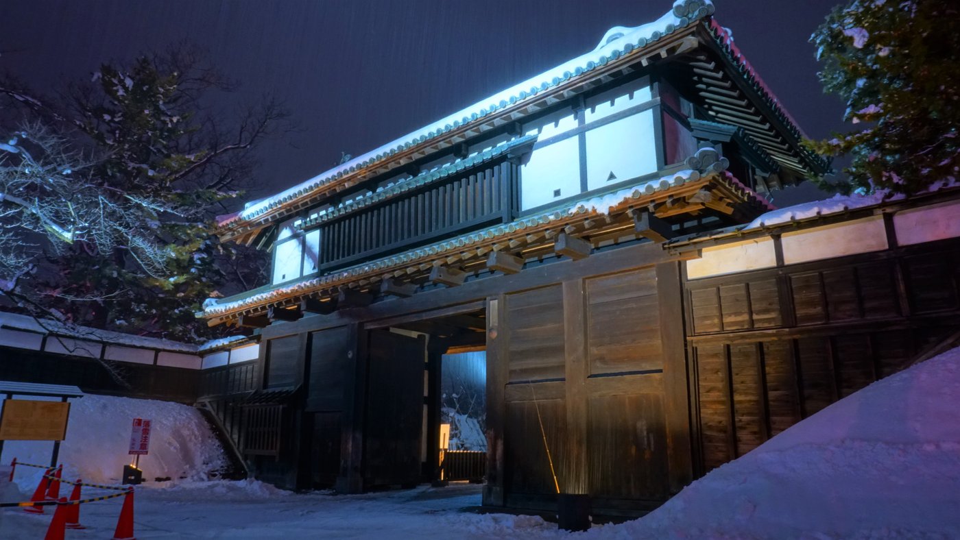 雪夜裡打燈的弘前城追手門,兩層木造城門屋頂積雪,門洞敞開,門前地面積雪,左側有三角錐與告示牌