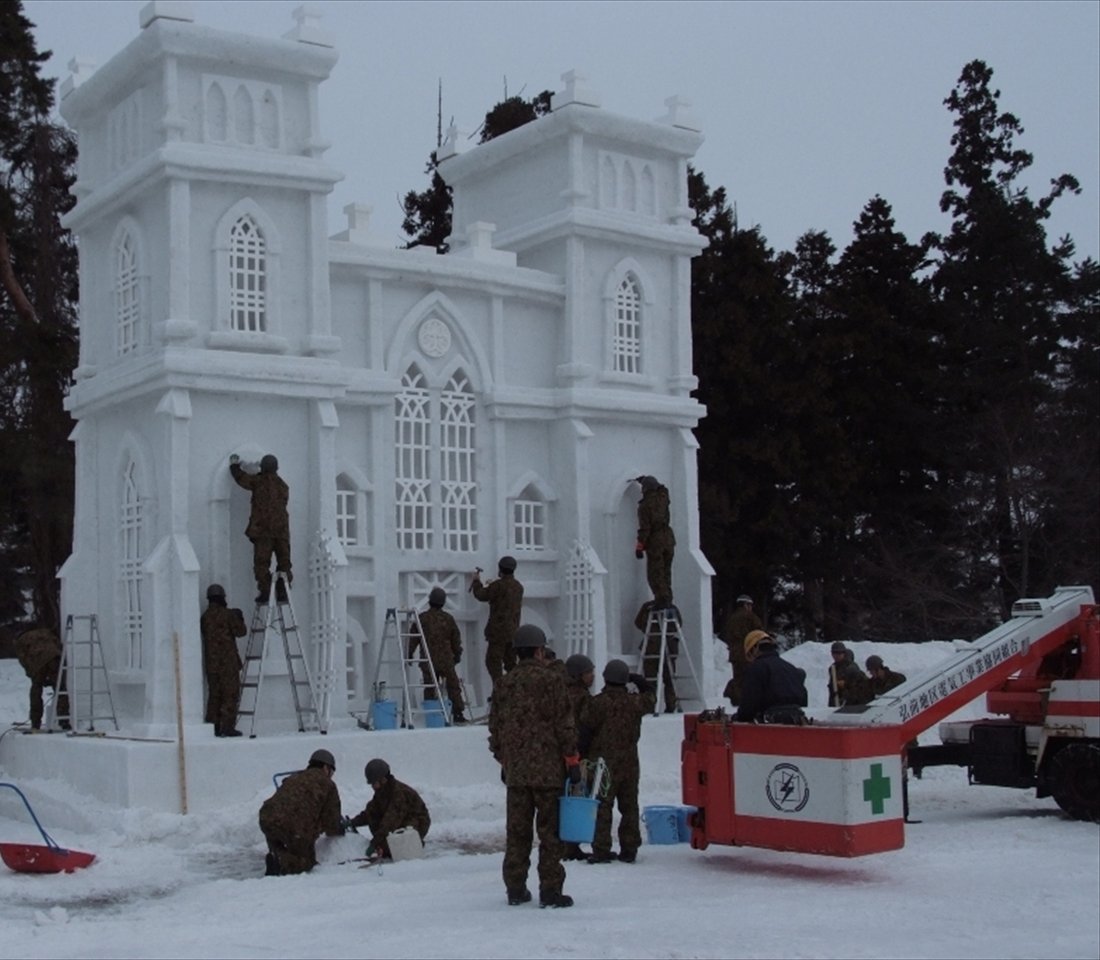 白天雪地上一座兩層樓高的哥德式教堂雪像,十多位穿迷彩服、戴頭盔的人站在鋁梯上雕窗戶細節,右邊停著一台高空作業車