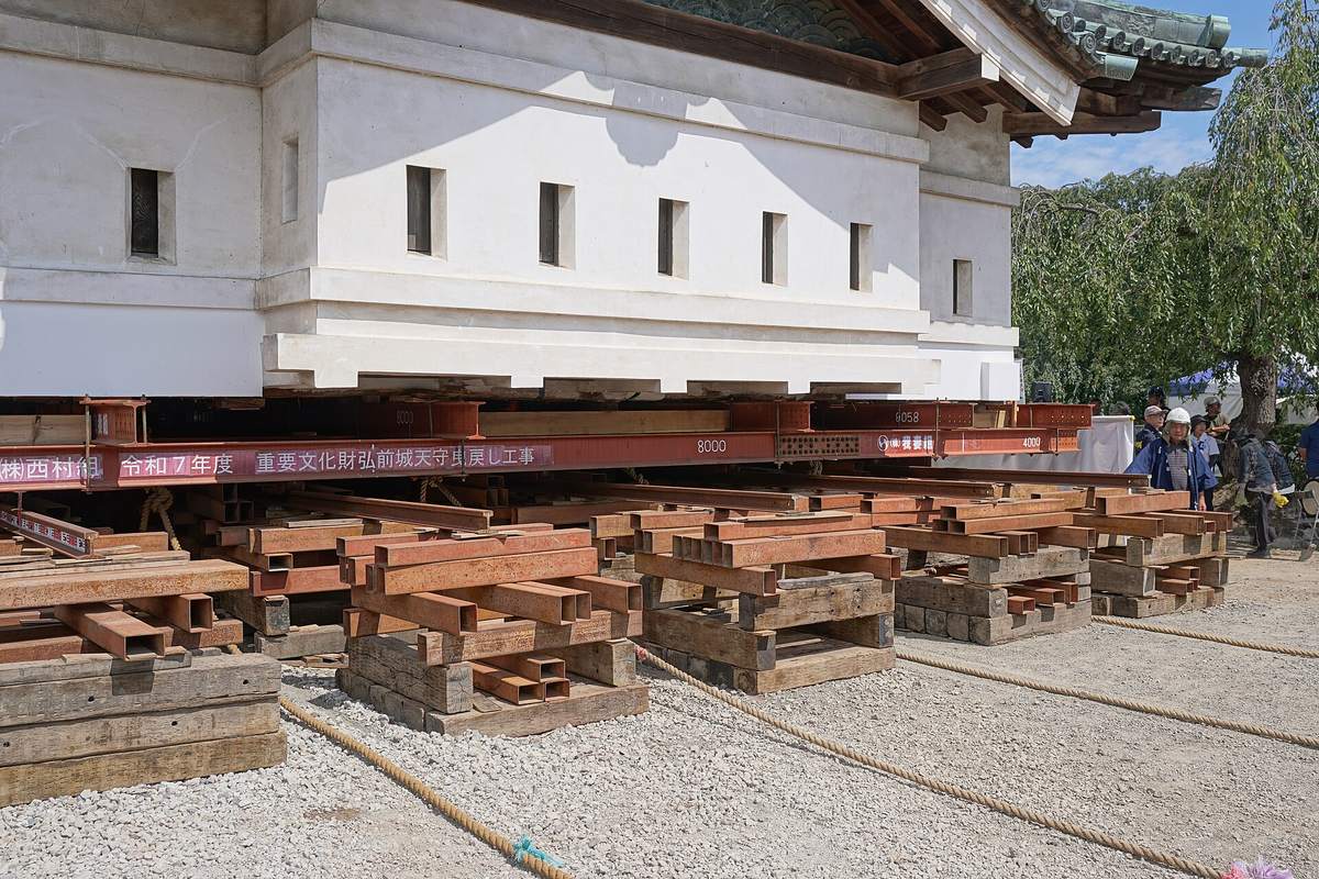 Close-up of the lower white wall of Hirosaki Castle keep resting on a row of red steel beams, with layers of interlocking timber cribbing and steel packing stacked beneath on gravel, thick straw ropes running across the ground and helmeted workers standing at the right