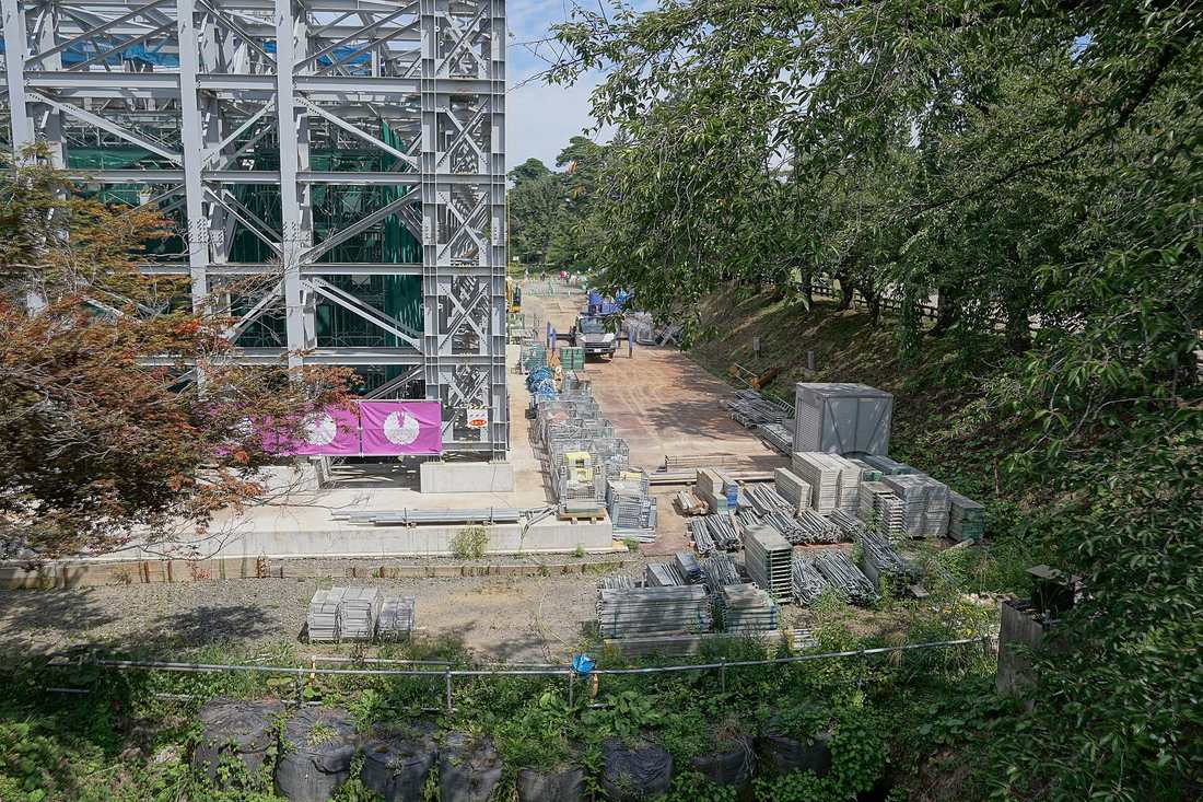 Looking down on the Hirosaki Castle inner moat work site: a grey steel lattice work platform rising from the dry moat floor, draped inside with green safety netting, standing on a concrete slab, with stacks of scaffolding boards and pipes on the bare earth to the right