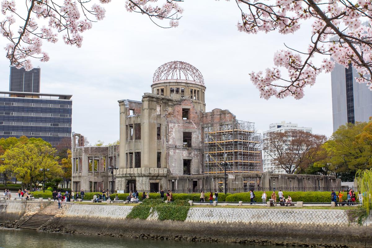 2016 年 4 月從平和記念公園的河岸隔著元安川看原爆圓頂,畫面上緣是河岸櫻花伸出來的枝條,圓頂右側搭著鷹架,對岸的步道與石階上有行人