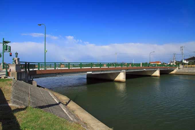 藍天下橫跨由良川的柯南大橋,綠色欄杆、橋頭有石柱與小銅像,從河岸草坡斜看過去,對岸遠處是低矮的住宅