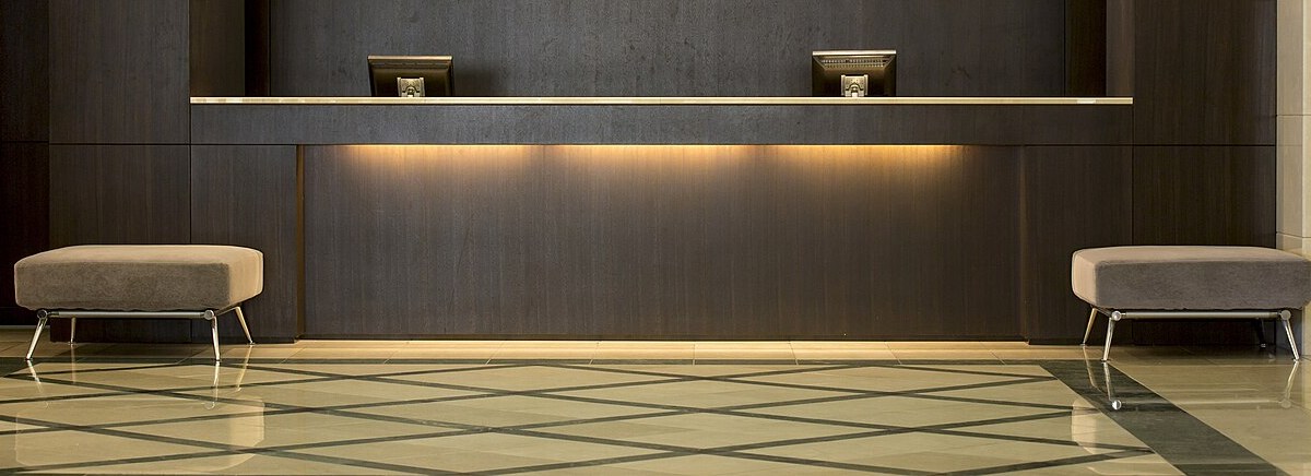 The long dark-wood front desk of a Japanese hotel lobby, two computer monitors facing the staff side, warm concealed lighting under the counter, a grey upholstered bench at each end and a cream-and-green diamond-patterned marble floor