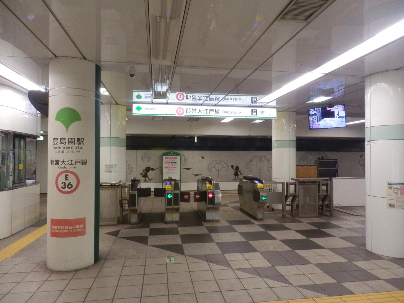 都營大江戶線豊島園站的地下剪票口：左邊白色圓柱上印著綠色銀杏標誌與「豊島園駅 Toshimaen Sta.」「都営大江戸線」「E 36」，中間一排自動剪票機，上方綠白色看板寫著「都営大江戸線 Oedo Line」，後方牆上有剪影圖案
