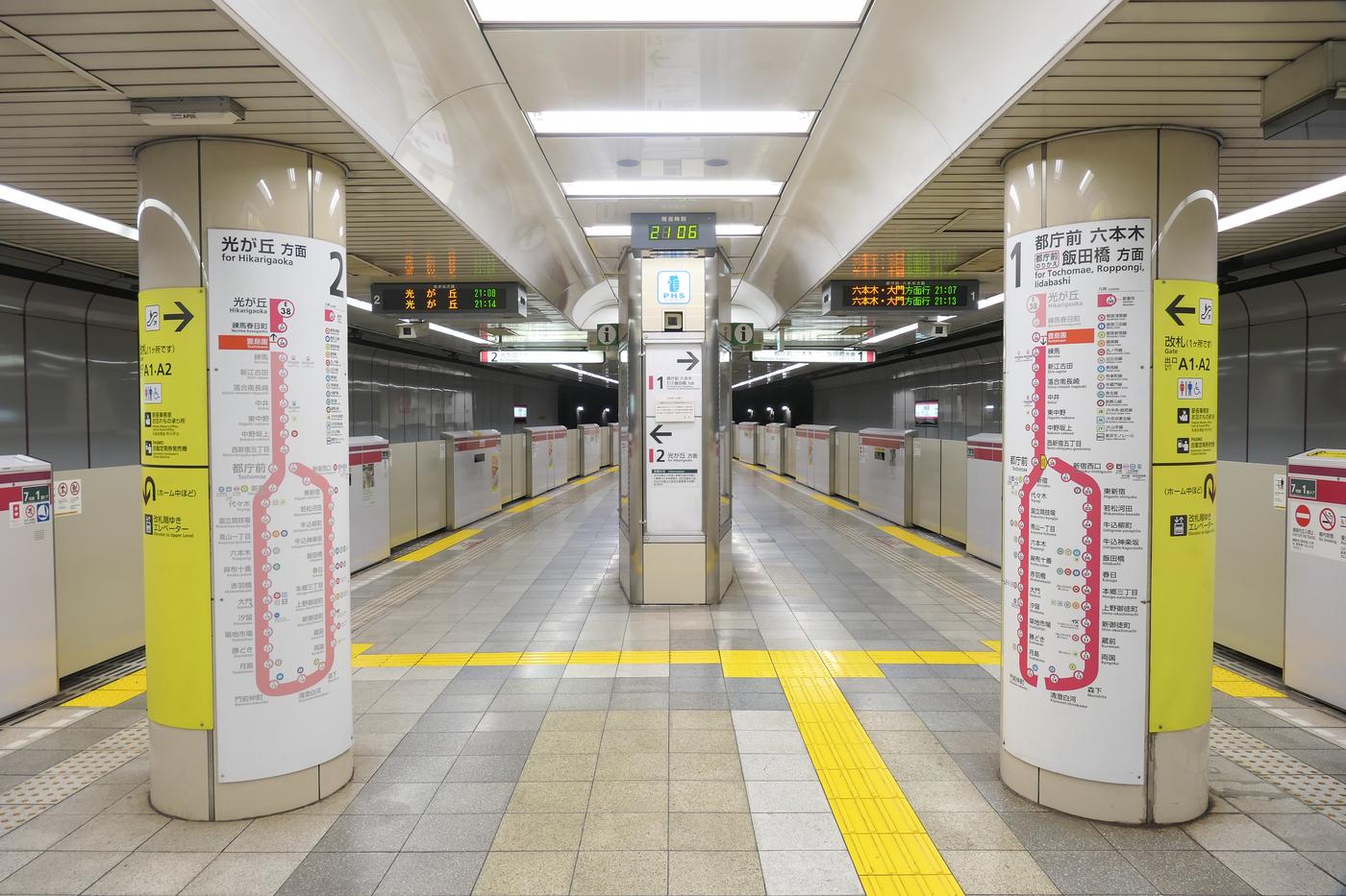 都營大江戶線豊島園站地下月台：左柱標示「2 光が丘方面」，右柱標示「1 都庁前 六本木 飯田橋方面」，兩柱貼著紅色路線圖與黃色出口指示，中間是月台門與一條黃色導盲磚，月台上沒有人