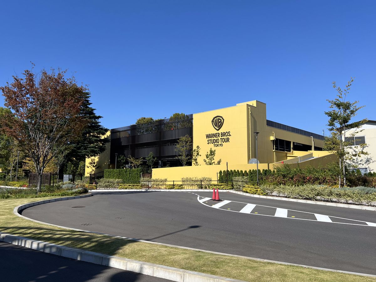 晴天下東京哈利波特影城的黃色大型建物，外牆上有華納兄弟盾牌標誌與「WARNER BROS. STUDIO TOUR TOKYO」字樣，前面是空的圓環車道與斑馬線，左側一棵開始轉紅的樹