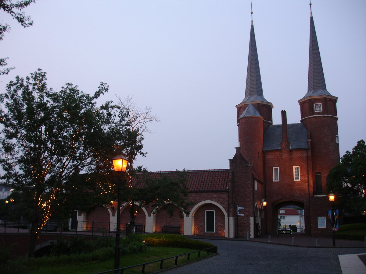 A red-brick twin-spired gatehouse and arcaded wall at Huis Ten Bosch at dusk, street lamps lit above a cobbled roadway
