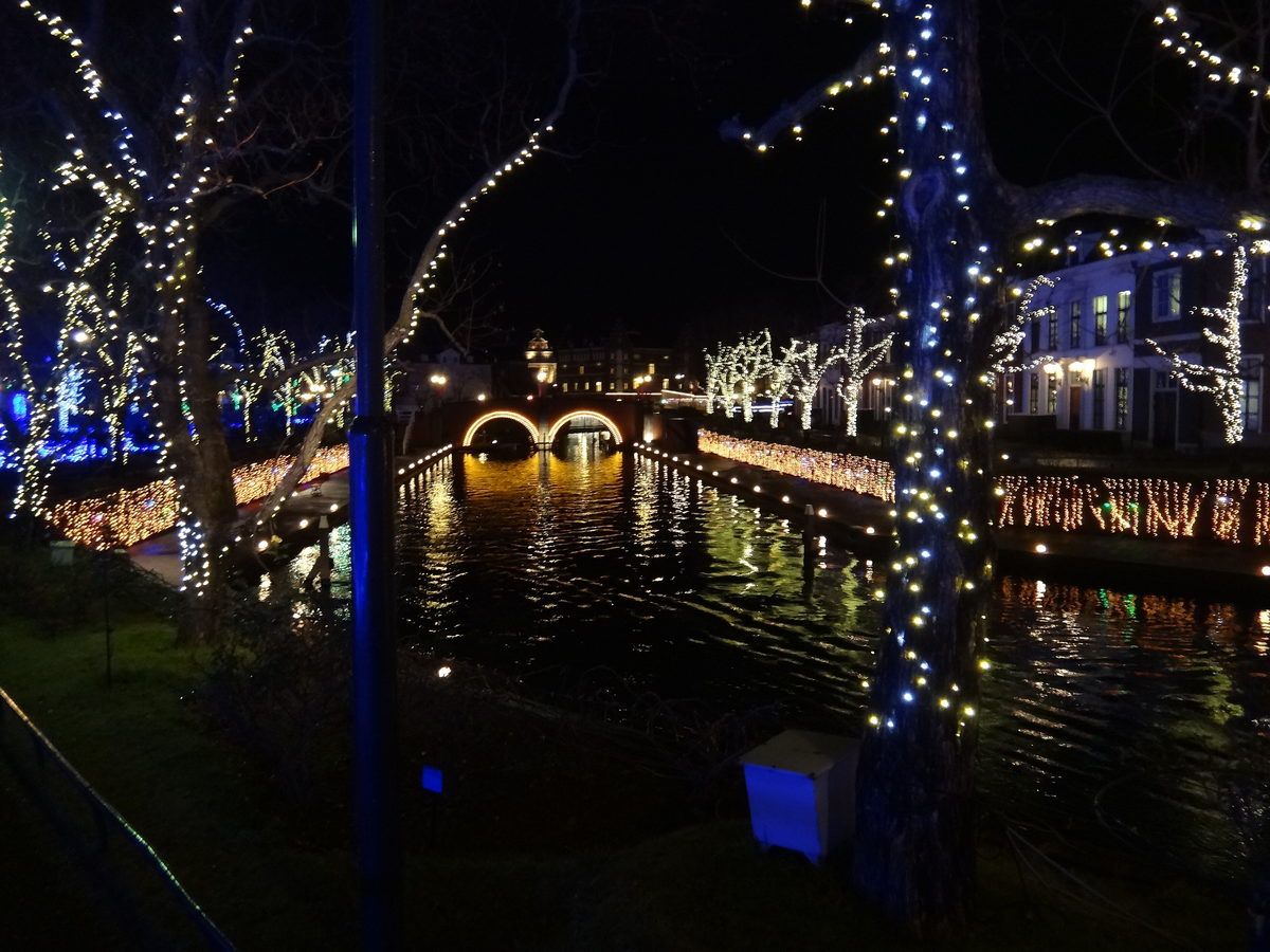 Night illumination at Huis Ten Bosch: trees wrapped in warm lights, curtain lights along the brick canal embankments and an arched bridge lit gold, all mirrored in the water