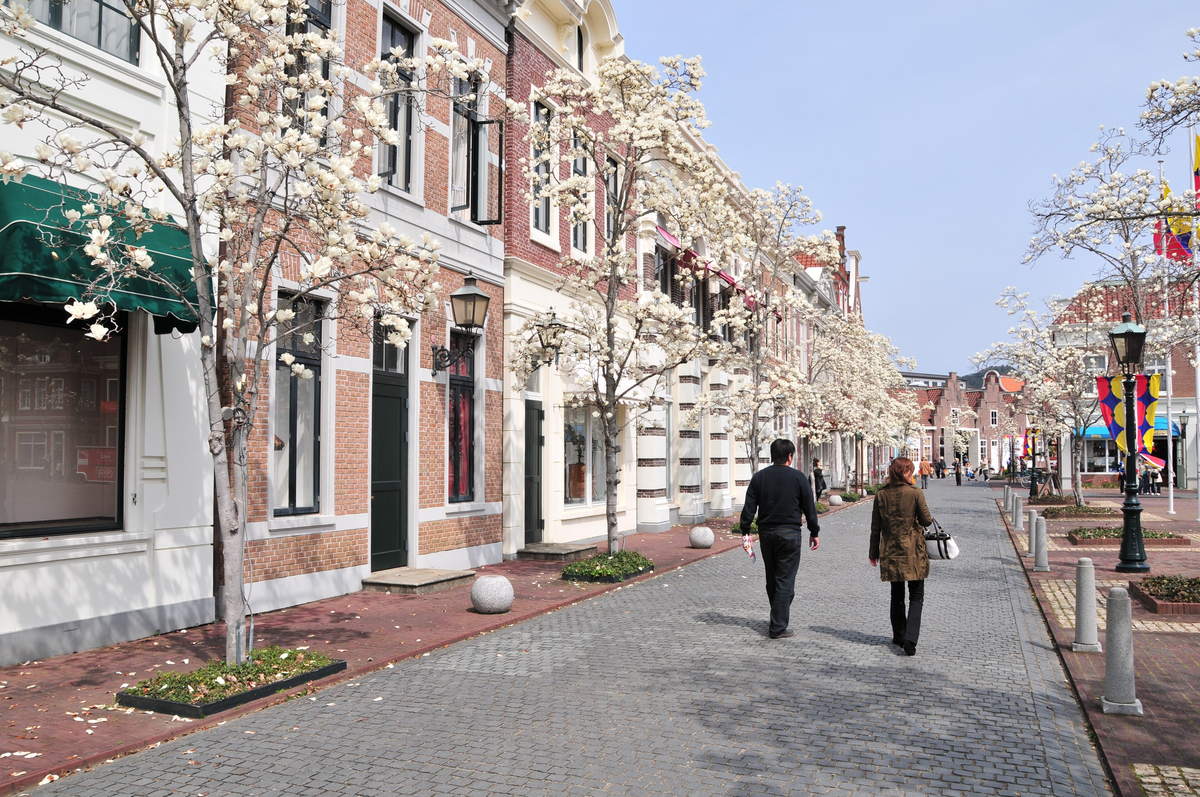 A cobbled shopping street inside Huis Ten Bosch lined with red-brick and white Dutch shopfronts, magnolia trees in white blossom, two visitors walking away from the camera