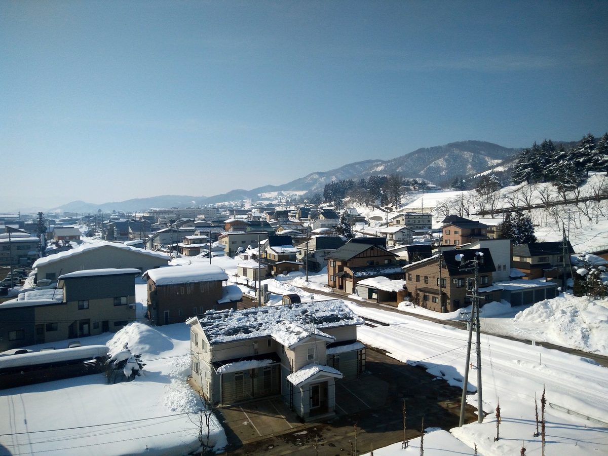 二月晴天從飯山站往外看的飯山市區:住宅屋頂積著厚雪,房子之間堆著剷出來的雪堆,右側那條路的路面整條是壓實的白雪與車轍,遠方是積雪的山稜