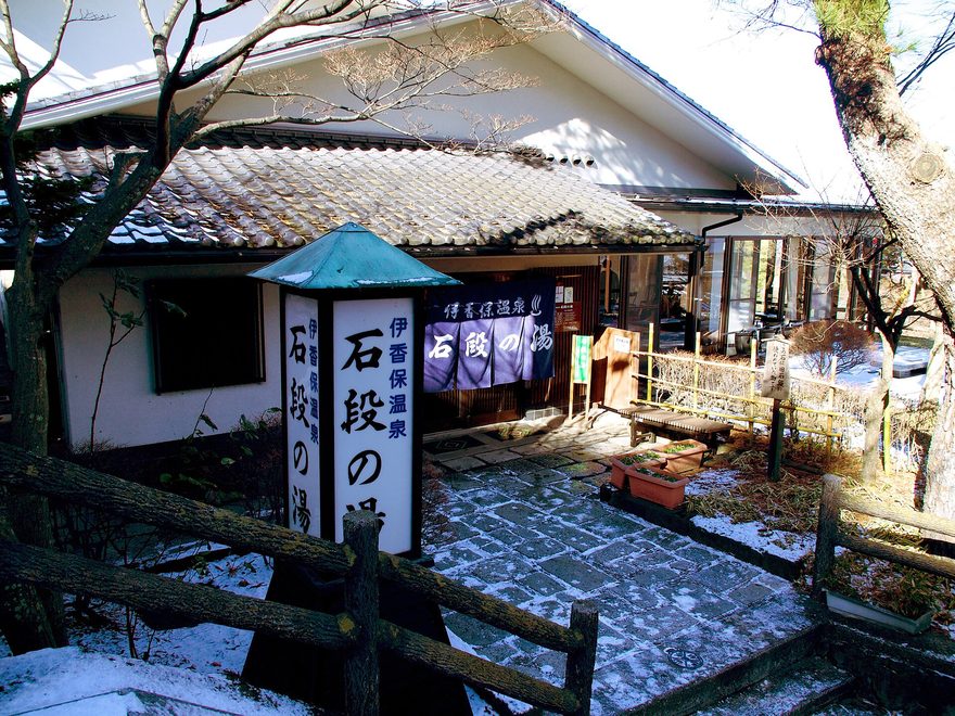 冬季的石段之湯外觀，瓦頂建築前立著印有「伊香保溫泉 石段之湯」的燈箱招牌與同名暖簾，石板地上有殘雪
