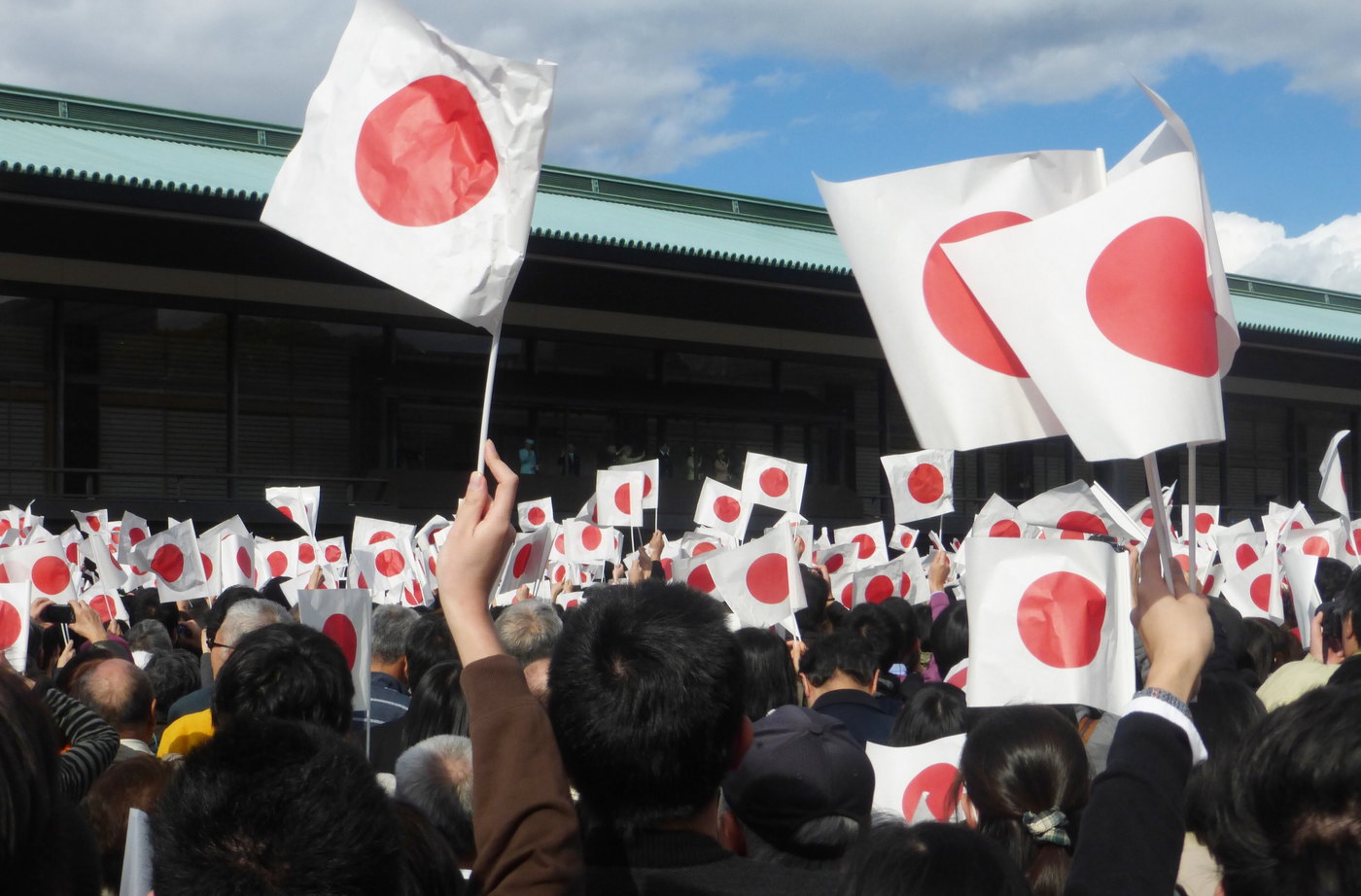 A crowd on the Imperial Palace east plaza holding up small paper Hinomaru flags towards the Chowaden Reception Hall, under a clear winter sky