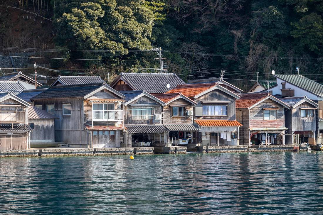 Ine funaya photographed from the water, ground-floor boat garages opening to the sea, upper floors with windows and balconies, stone bases dropping straight into the bay
