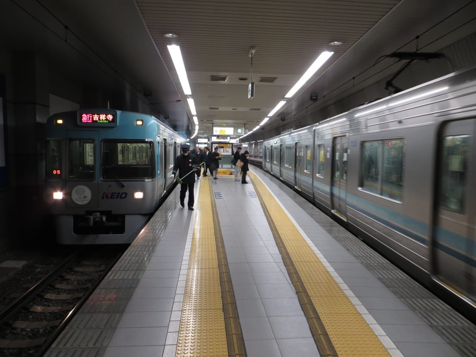 A Keio Corporation train standing at an enclosed underground platform, KEIO lettering below the cab window and a red LED destination panel above it reading Express Kichijoji in Japanese and English, a uniformed station attendant walking down the platform with a long-handled tool, and a second train passing on the right